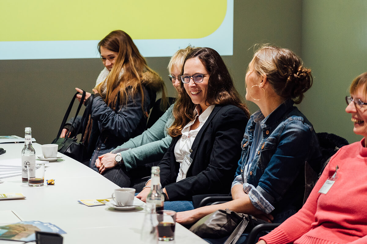 Mehrere lachende Frauen sitzen an einem Besprechungstisch. Im Hintergrund ist eine Präsentation per Beamer an die Wand geworfen.