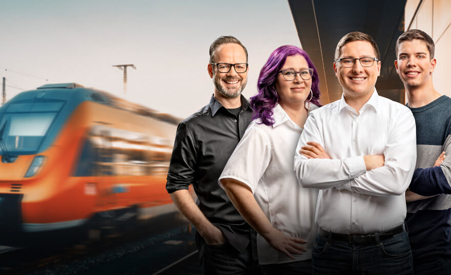Drei Lokführer und eine Lokführerin posieren gemeinsam und lächeln in die Kamera. Im Hintergrund ist ein Zug.