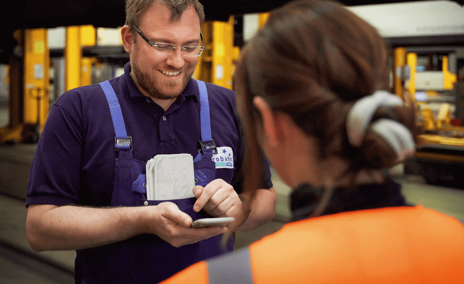 Ein Werkstattmitarbeiter schaut auf sein Handy und lächelt, im Vordergrund ist eine Frau zu sehen, die auf sein Handy schaut.