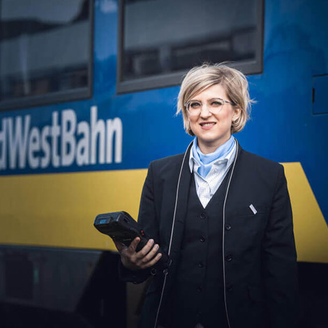 Blonde, lächelnde Frau steht in einer Bahnuniform mit einem Scanngerät vor einer blau-gelben Bahn.