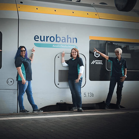Drei Frauen stehen vor einer S-Bahn, auf der Eurobahn steht, zeigen auf diesen Schriftzug und lächeln in die Kamera.
