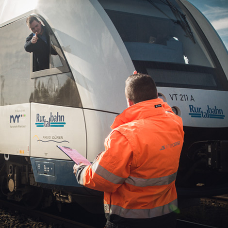 Ein Mann mit oragener Warnjacke steht vor einer Bahn. Sein Rücken ist zur Kamera gerichtet.