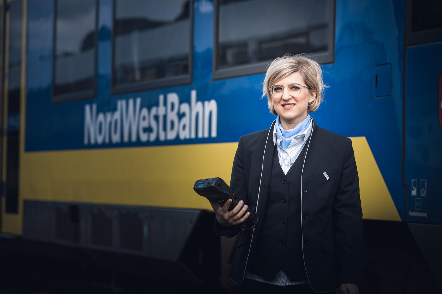 Frau in Uniform, Halstuch und einem Ticketgerät in der Hand steht vor einer blauen Regionalbahn, auf der NordWestBahn zu lesen ist.