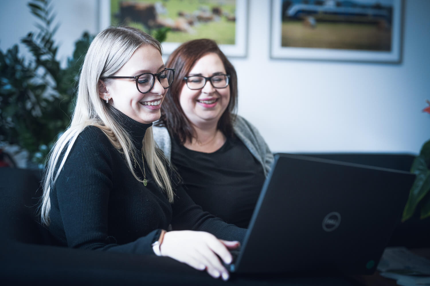 Zwei junge Frauen sitzen gutgelaunt in einem Raum und schauen beide auf einen aufgeklappten Laptop.