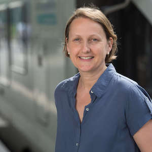 Portraitfoto von Anne Mathieu: Dunkelblonde Frau in blauer Bluse vor einem Zug