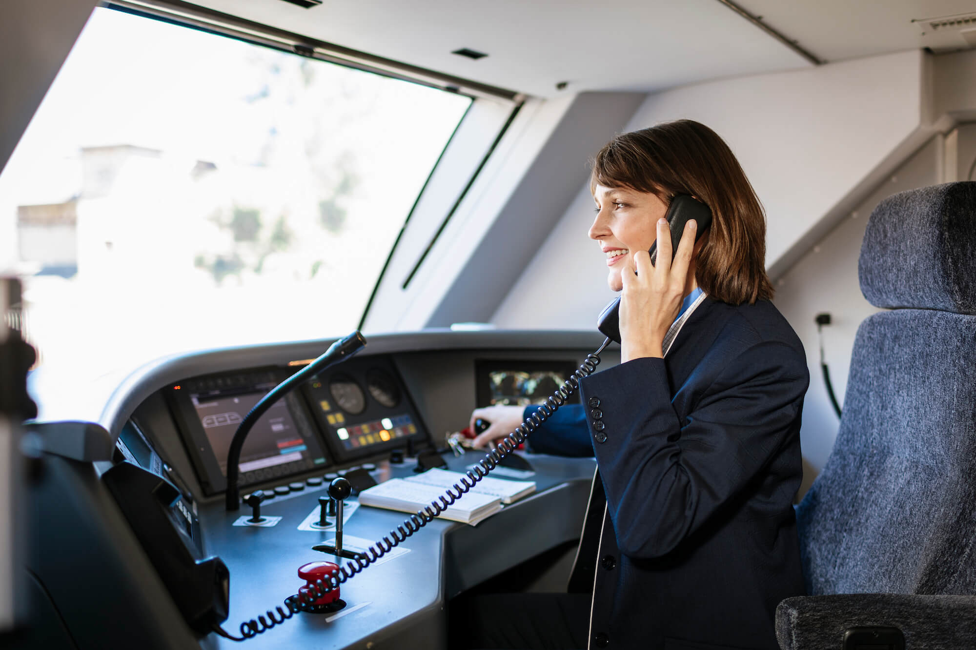 Lokführerin sitzt im Führerhaus einer Bahn, man sieht den Techniktisch mit vielen Schaltern und Knöpfen. Die Frau telefoniert mit dem Boardtelefon.