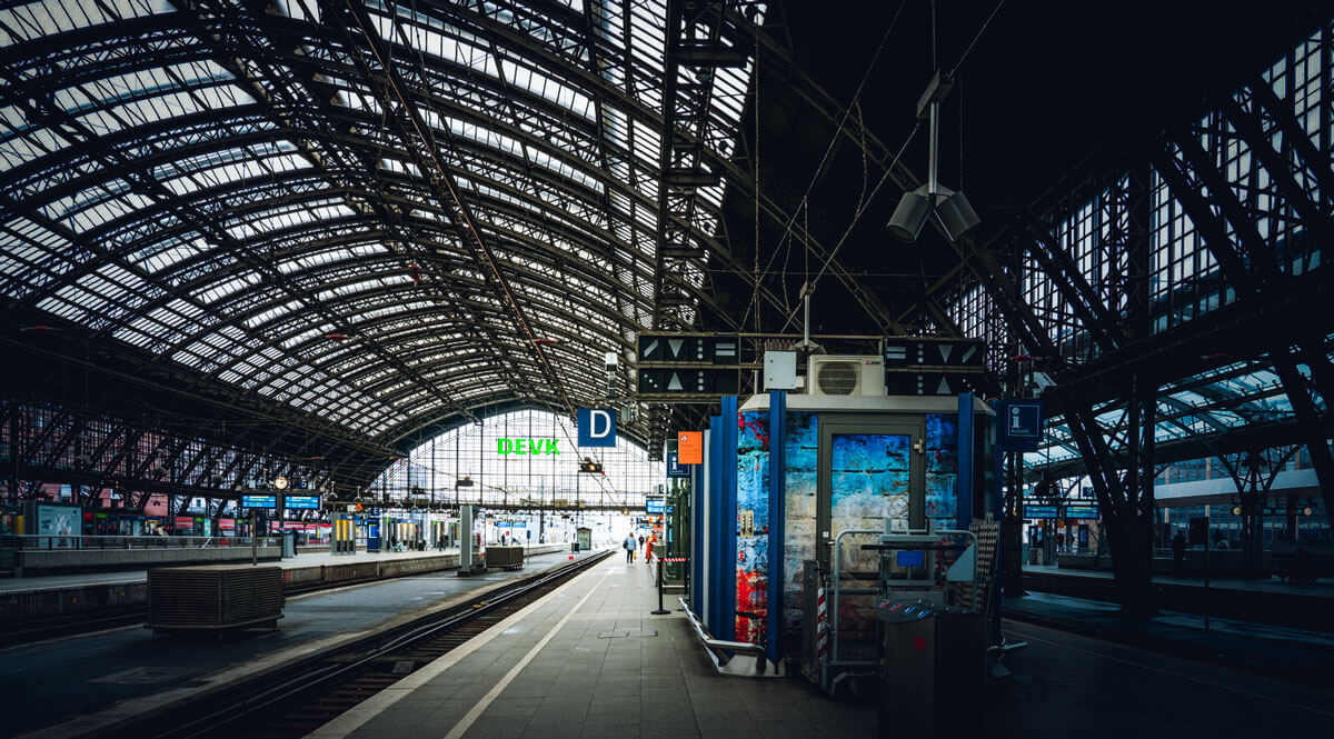 In der Ferne fällt licht auf ein Gleis im Kölner Hauptbahnhof. Das Gleis wird  von einem Dach aus Stahlstreben überdacht, um Vordergrund ist ein Aufsichtshäuschen zu sehen.