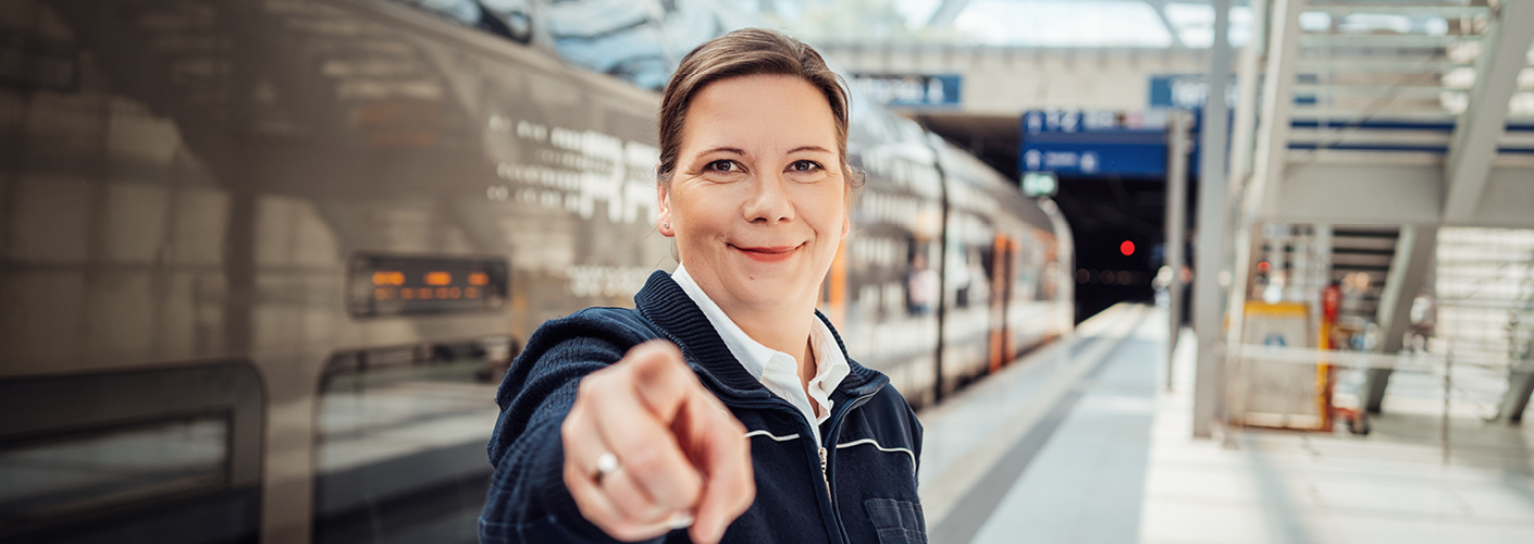 Eine dunkelblonde Frau in Uniform steht vor einem RRX-Zug am Bahnsteig und zeigt auf etwas Besonderes.