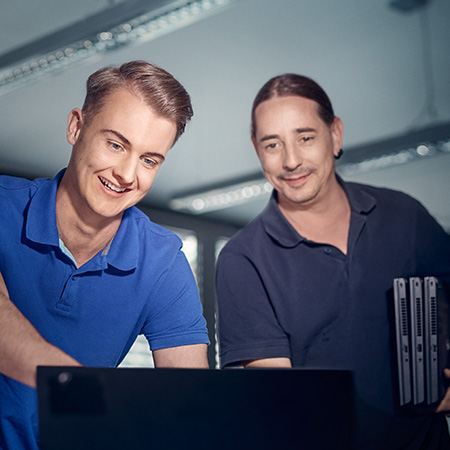Ein IT-Spezialist schaut mit seinem Kollegen auf einen Bildschirm. Er lächelt und trägt ein blaues Polo-Shirt.