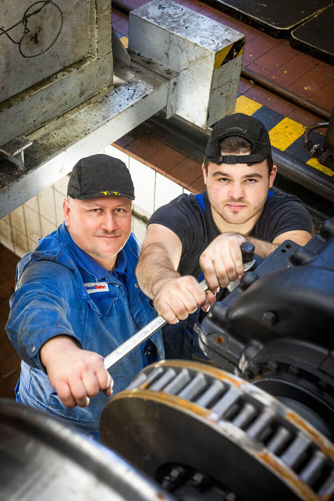 Zwei Männer in Handwerker-Outfit schauen in die Kamera während sie Werkzeug in der Hand halten und in einer Werkstattgrube stehen. 