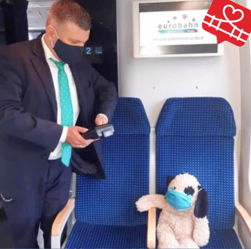 Ein Kundenbetreuer und ein Stofftier in einer Bahn mit jeweils einer Atemschutzmaske im Gesicht.