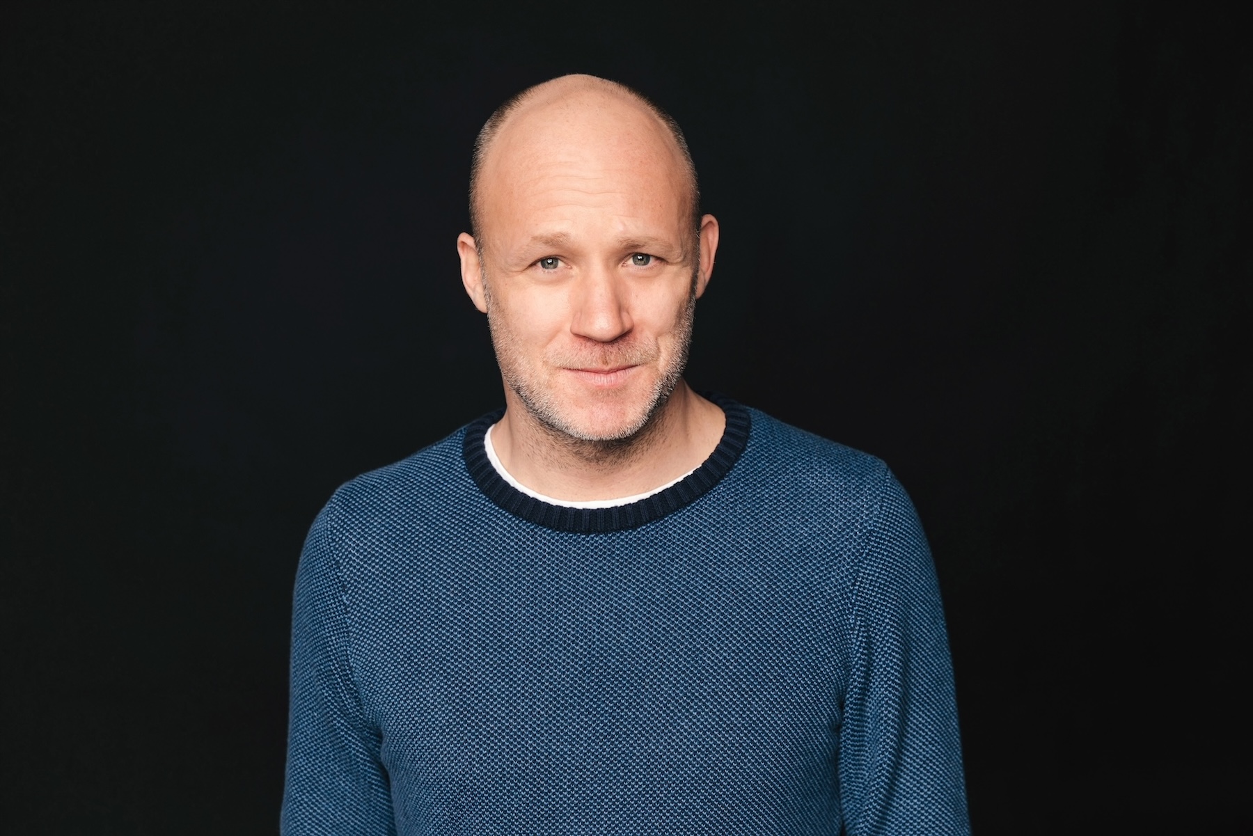 Moderator Andreas Bursche steht vor einem dunklen Hintergrund. 