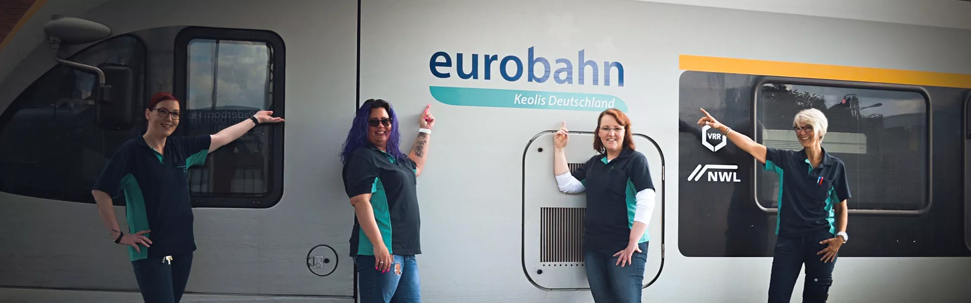 Vier Frauen stehen gut gelaunt vor einem weißen Zug. auf dem eurobahn steht und zeigen mit den Händen auf ebendiesen Schriftzug.