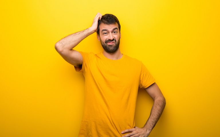 Ein Mann in einem gelben T-Shirt steht vor einer gelben Wand und reibt sich verwirrt den Kopf.