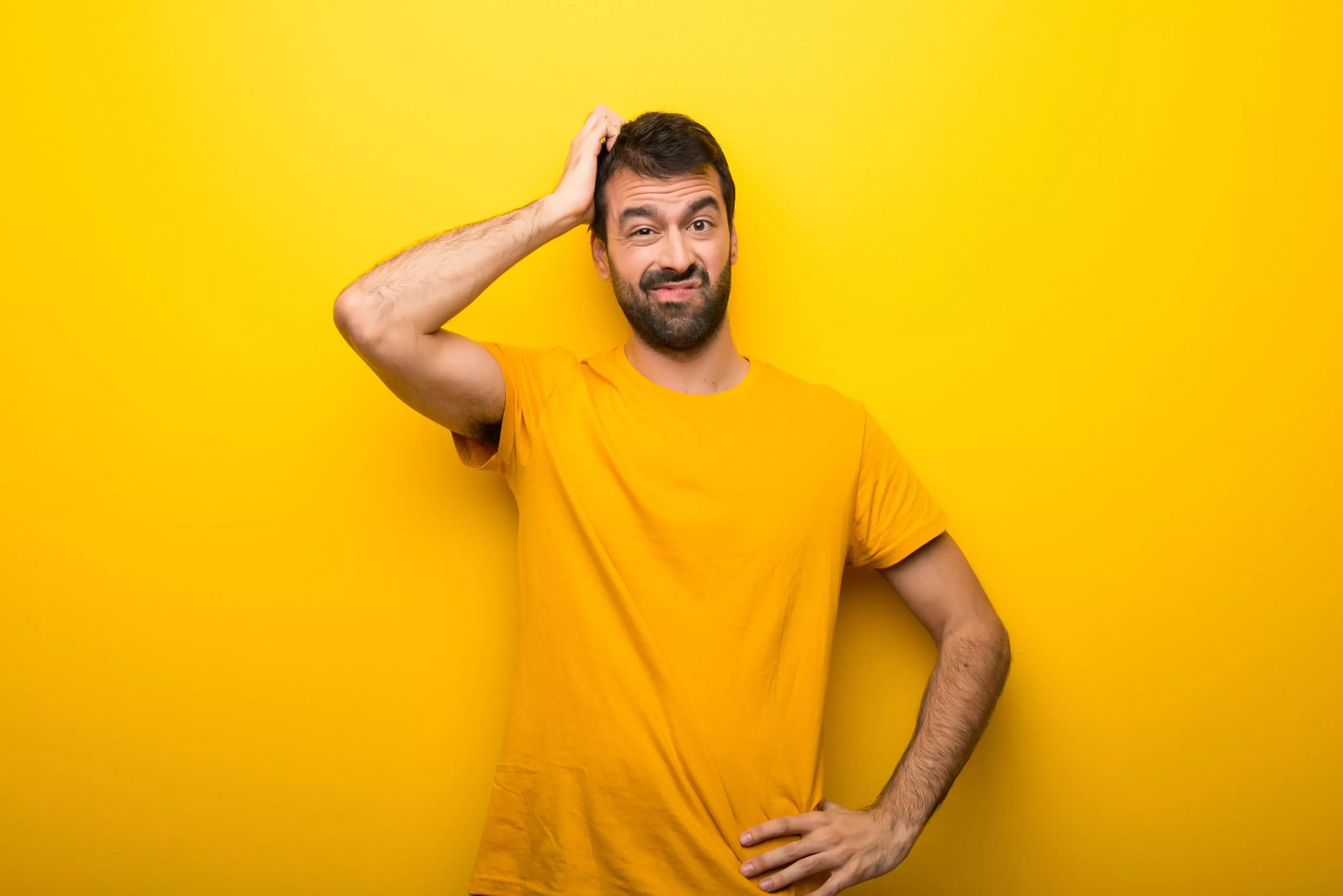 Ein Mann in einem gelben T-Shirt steht vor einer gelben Wand und reibt sich verwirrt den Kopf.