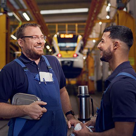 Zwei Werkstattmitarbeiter schauen sich an und lachen. Sie halten eine Brotdose und eine Thermoskanne, im HIntergrund ist ein Zug auf einer Heberampe zu sehen.