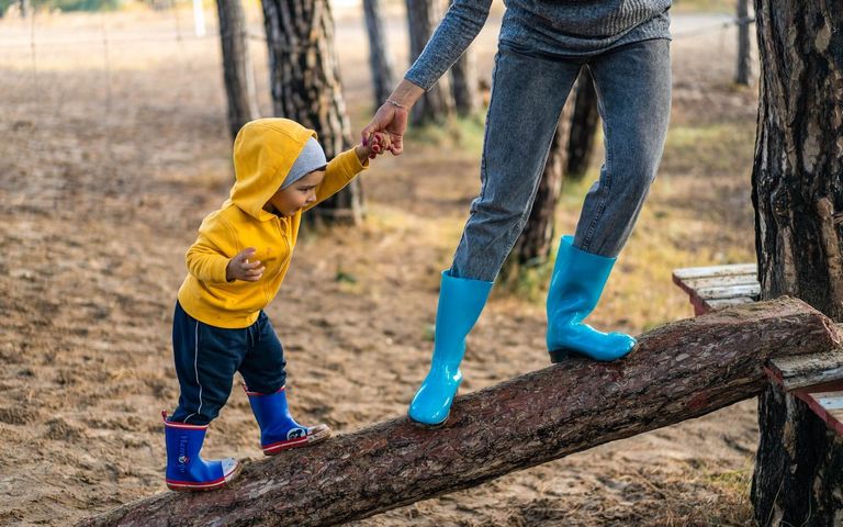 Ein kleiner Junge mit gelber Regenjacke klettert an der Hand seiner Mutter auf einem Baumstamm. 