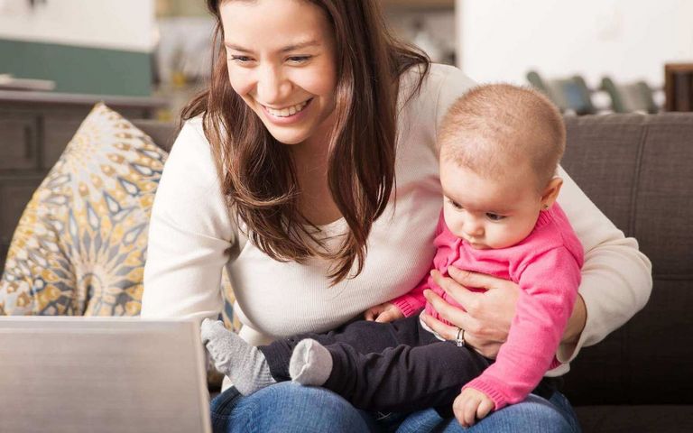 Eine Frau sitzt mit ihrem Baby im Arm auf der Couch und arbeitet am Laptop