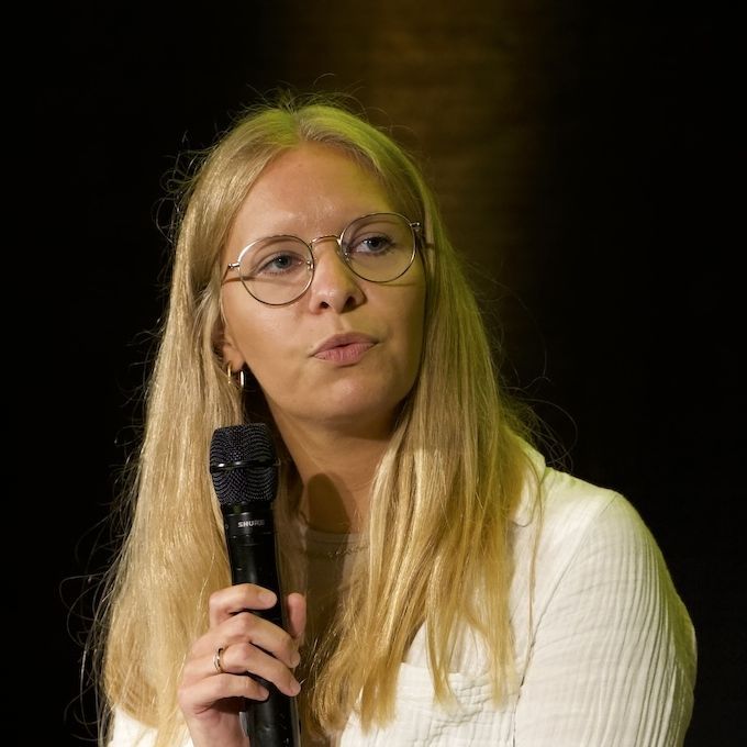 Eine blonde, junge Frau mit Brille sitzt in einem Sessel mit einem Mikrofon in der Hand.