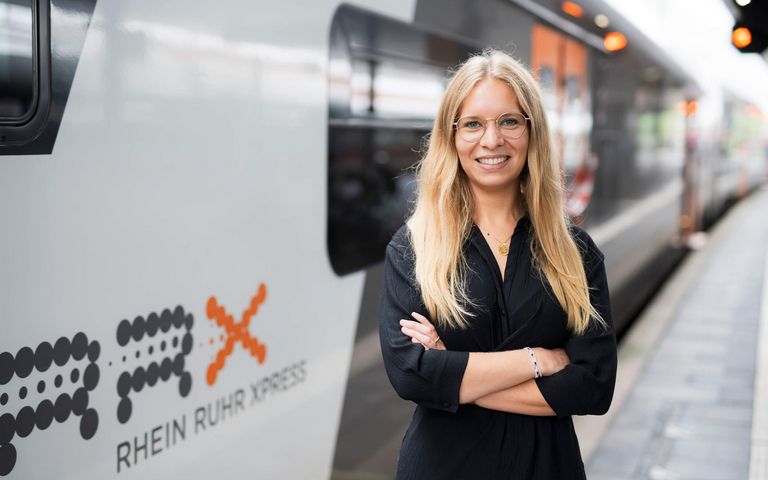 Eine blonde Frau mit Brille steht vor einem RRX-Zug und lächelt in die Kamera.