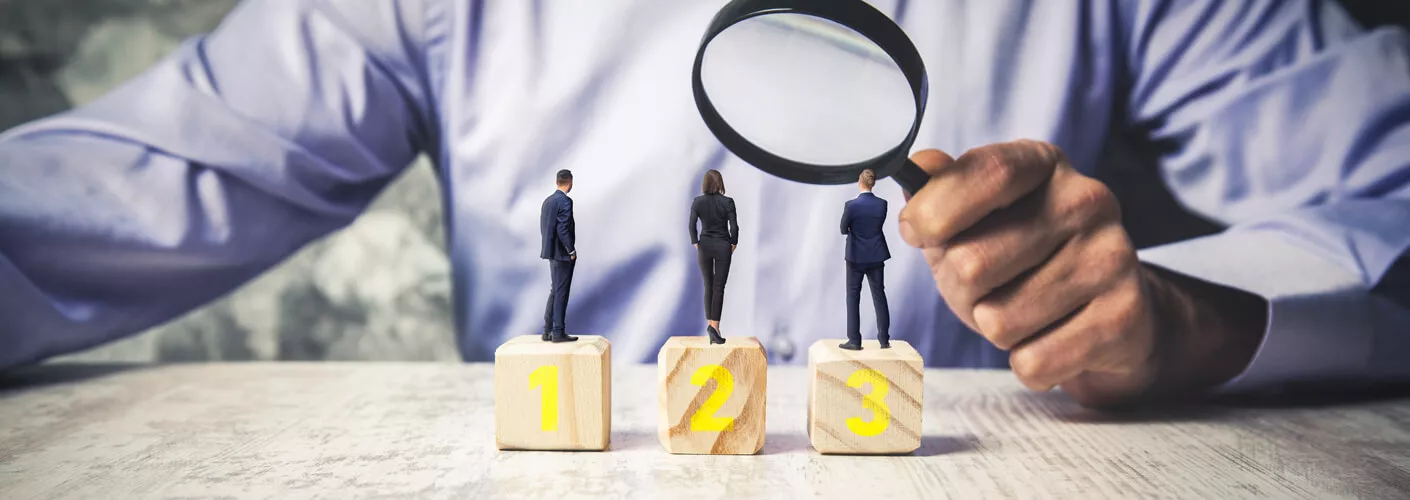 Anischt eines Mannes in Businesskleidung der durch eine Lupe drei Figuren betrachtet. Diese sind als ebenfalls in Businesskleidung und stehen auf drei Holzblöcken auf denen die Zahlen 1 bis 3 stehen.