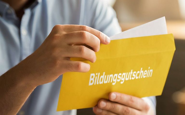 Ein Mann hält einen gelben Umschlang in der Hand und öffnet ihn. Auf dem Umschlag ist in weißen Buchstaben das Wort "Bildungsgutschein" zu lesen.