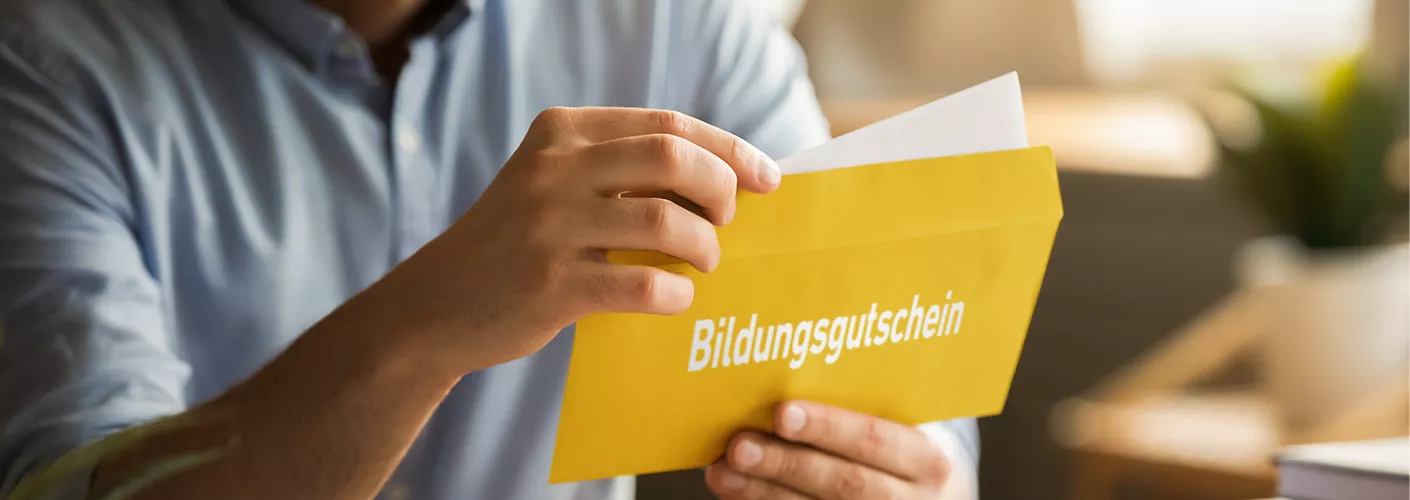 Ein Mann hält einen gelben Umschlang in der Hand und öffnet ihn. Auf dem Umschlag ist in weißen Buchstaben das Wort "Bildungsgutschein" zu lesen.