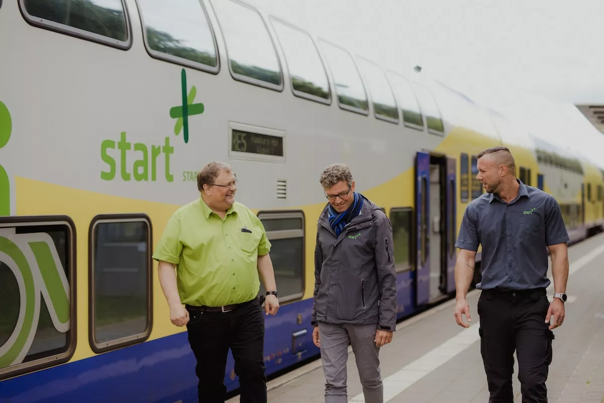 Ein Zug des Unternehmens Start Deutschland steht an einem Bahnhof, drei Mitarbeitende laufen davor herum. 