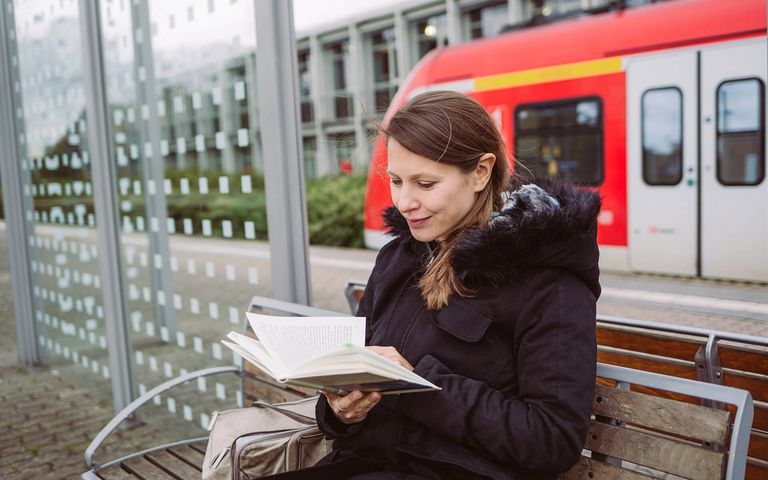 Sarah Meyer-Dietrich sitzt an einem Bahnsteig in einem Wartehäuschen und liest ein Buch.