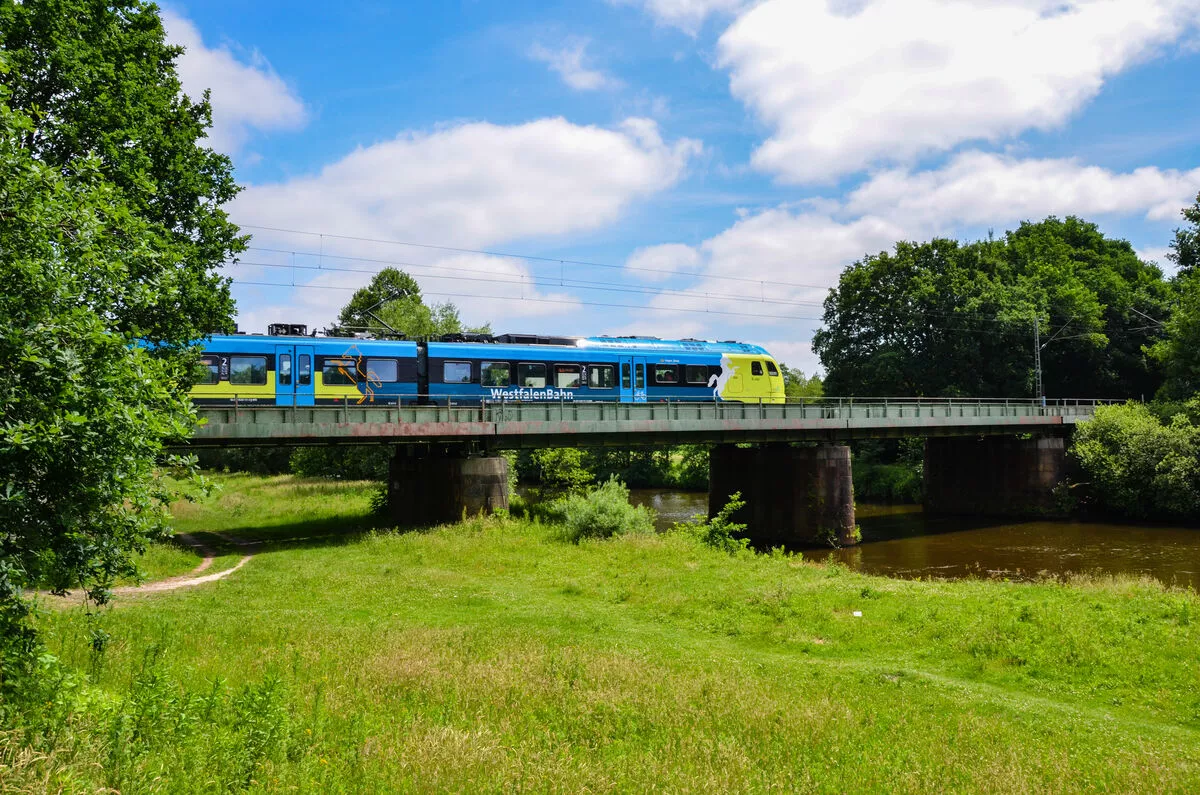Ein blauer Zug fährt über eine Brücke, die Sonne scheint auf grünes Gras.