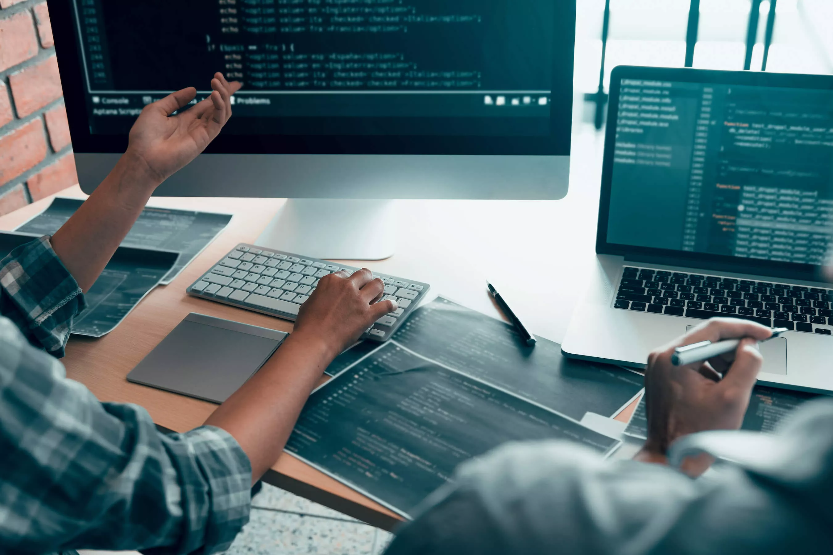 Rückansicht zweier Personen, die vor einem PC und Laptop sitzen und darauf programmieren.