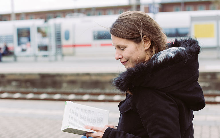 Sarah Meyer-Dietrich sitzt an einem Bahnsteig und liest ein Buch.