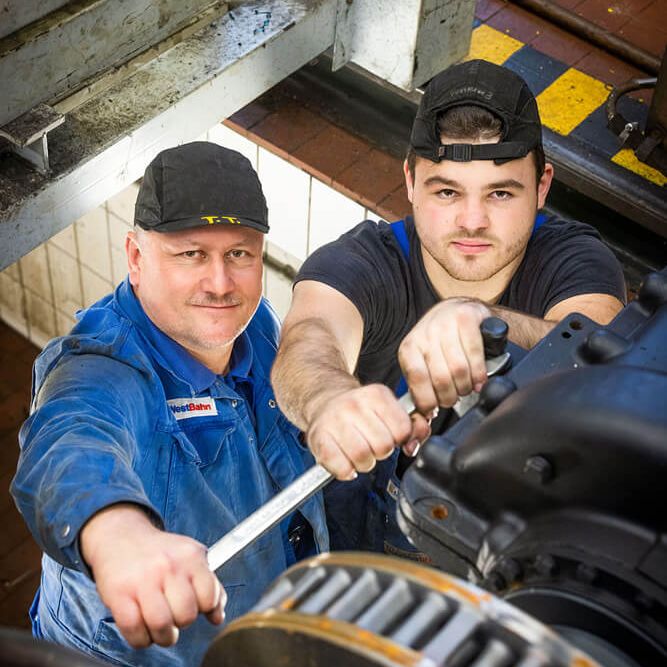 Zwei Männer in Handwerker-Outfit schauen in die Kamera während sie Werkzeug in der Hand halten und in einer Werkstattgrube stehen. 