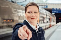 Triebfahrzeugführerin Claudia Stefanie Eine dunkelblonde Frau in Uniform steht vor einem RRX-Zug am Bahnsteig und zeigt auf etwas Besonderes.