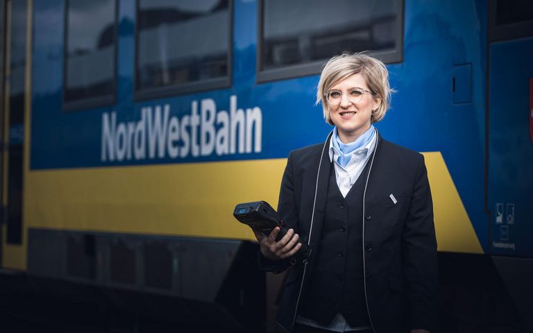 Frau in Uniform, Halstuch und einem Ticketgerät in der Hand steht vor einer blauen Regionalbahn, auf der NordWestBahn zu lesen ist.