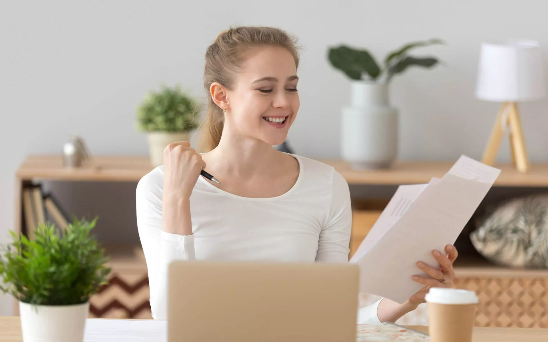 Eine junge Frau sitzt mit Papieren in der Hand am Laptop und freut sich.