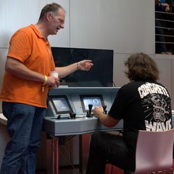 Ein Mann in orangenem Polo zeigt einer jungen Person im schwarzen Shirt ein Bildschirm mit Bedienelementen.