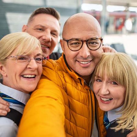 Ein Mann in gelber Jacke mit Schwarzer Brille macht ein Selfie von sich und seinen Kolleg*innen: Sie lächeln alle gemeinsam in die Kamera.