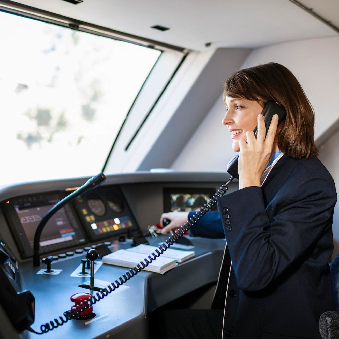 Lokführerin sitzt im Führerhaus einer Bahn, man sieht den Techniktisch mit vielen Schaltern und Knöpfen. Die Frau telefoniert mit dem Boardtelefon.