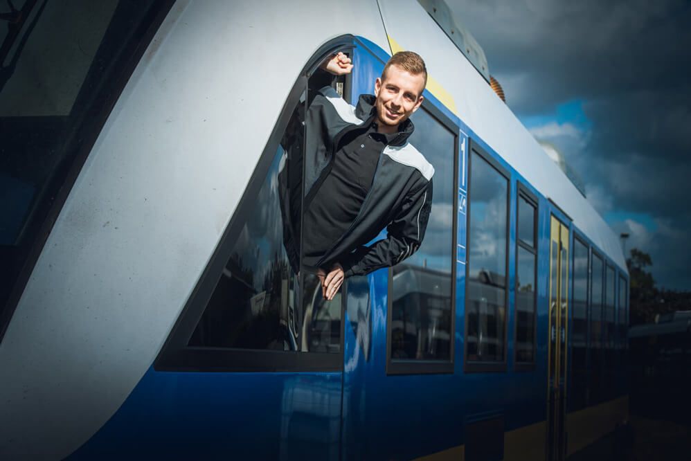 Mann in Lokführer-Outfit lehnt sich aus dem Fenster einer blauen Regionalbahn und schaut lächelnd in die Kamera.