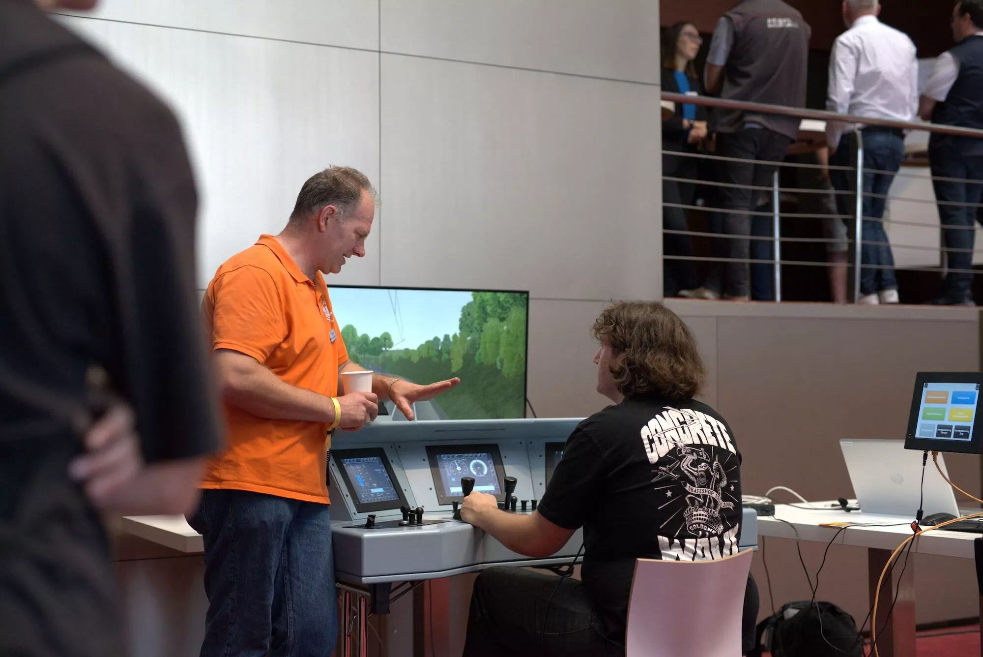 Ein Mann in orangenem Polo zeigt einer jungen Person im schwarzen Shirt ein Bildschirm mit Bedienelementen.