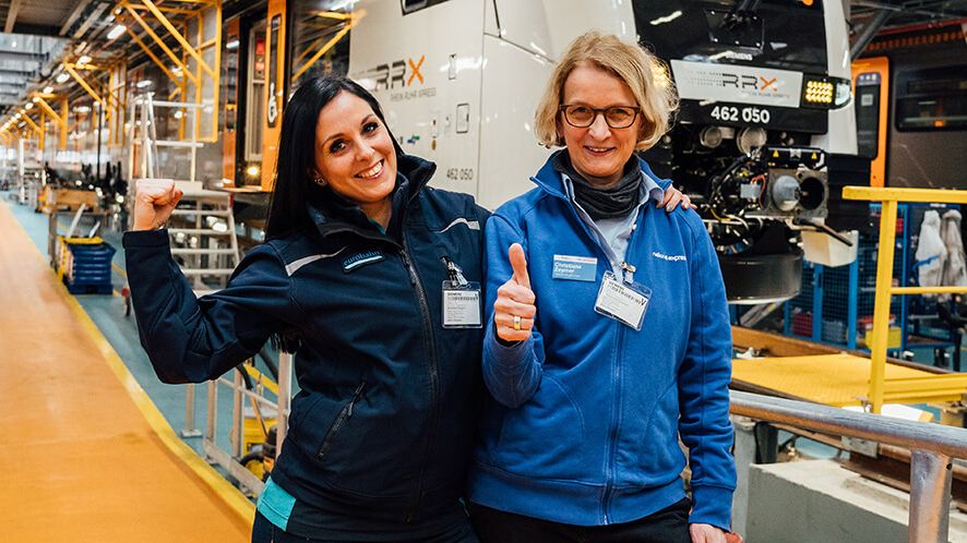 Zwei Frauen stehen auf Siemens-Werkgelände vor einem RRX-Zug. Die eine Frau lächelt und streckt den Daumen hoch, die andere flext ihren Oberarm.