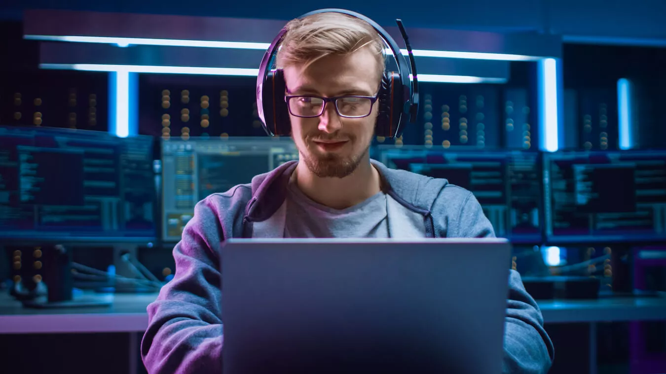 Ein Mann mit einem Headset auf dem Kopf sitzt fokussiert vor eiem Laptop. Im Hintergrund sind weitere Bildschirme zu sehen. 