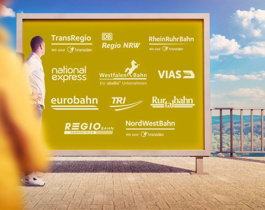 Frau im Vorder- und Mann im Hintergrund stehen vor einer goldfarbig unterlegten Tafel, auf der verschiedene Logos der Bahnunternehmen in NRW in weißer Farbe stehen.