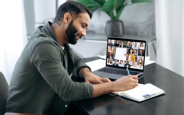 Ein Mann sitzt mit einem Notizbuch an einem Schreibtisch, auf seinem Laptop findet eine Videokonferenz statt.