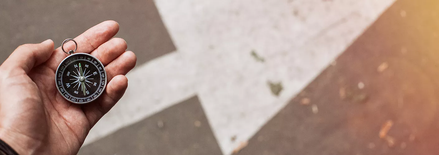 Eine Person hält einen Kompass in der Hand. Auf dem Boden ist ein Pfeil zu sehen, der nach links-oben zeigt.