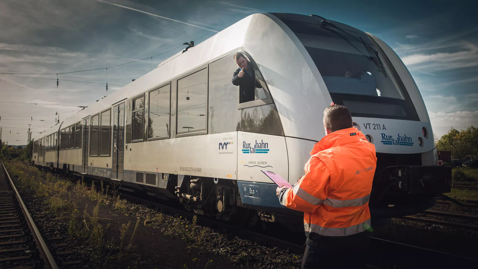 Ein Arbeiter mit orangener Warnjacke steht vor einem Zug, auf dem Rurtalbahn zu lesen ist, und zeigt dem Lokführer Daumen hoch. Der Lokführer schaut aus dem Cockpit-Fenster und erwidert den Daumen.