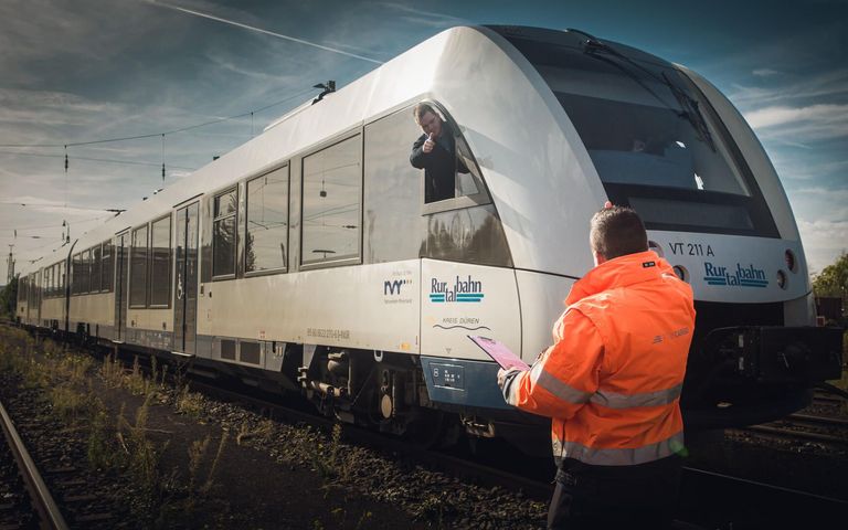 Ein Arbeiter mit orangener Warnjacke steht vor einem Zug, auf dem Rurtalbahn zu lesen ist, und zeigt dem Lokführer Daumen hoch. Der Lokführer schaut aus dem Cockpit-Fenster und erwidert den Daumen.
