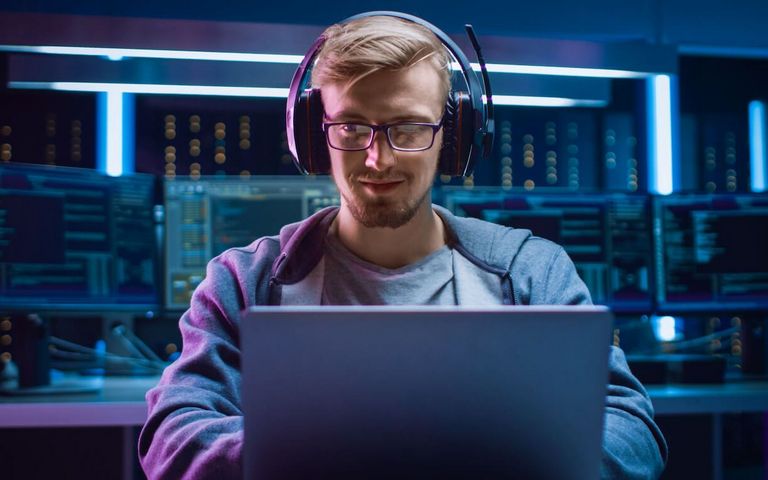 Ein Mann mit einem Headset auf dem Kopf sitzt fokussiert vor eiem Laptop. Im Hintergrund sind weitere Bildschirme zu sehen. 