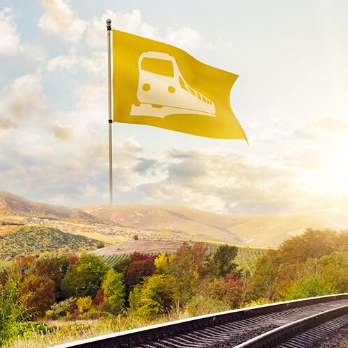 Panoramaaufnahme eines Gleises, dass durch eine flache Landschaft führt. Neben dem Gleis steht eine gelbe Flagge mit einem Zug-Symbol drauf.